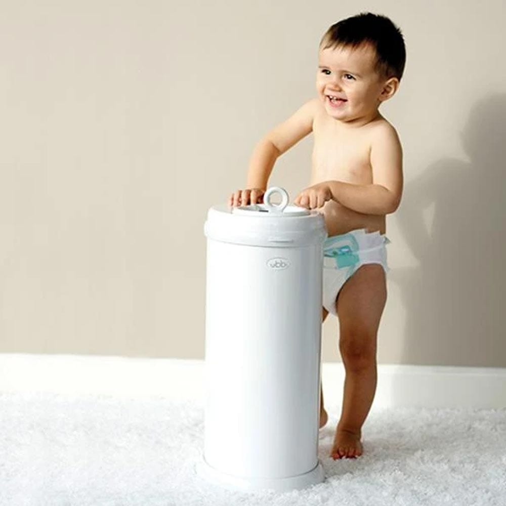 A baby standing by the diaper pail by Ubbi.