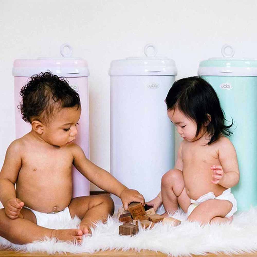 Two babies playing with blocks on a fur rug in from of three different colored Ubbi Diaper Pails. 